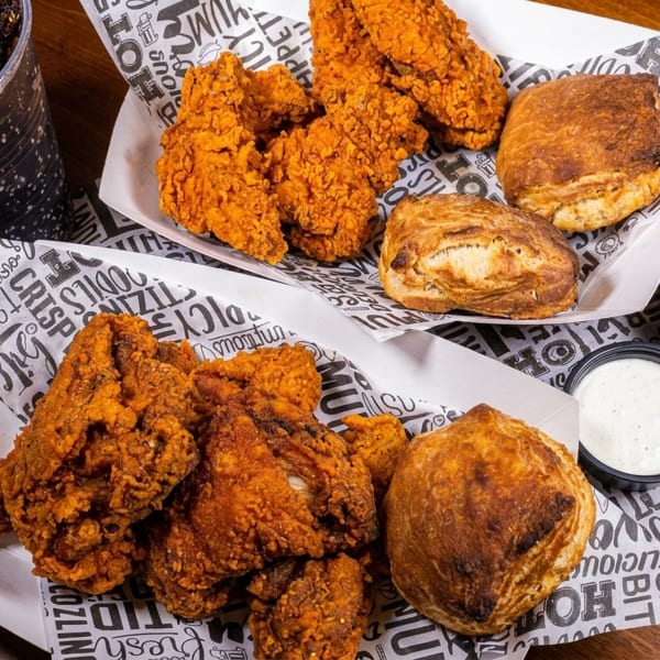 Two baskets of fried chicken and biscuits, with a soda and three dipping sauces on a wooden table.