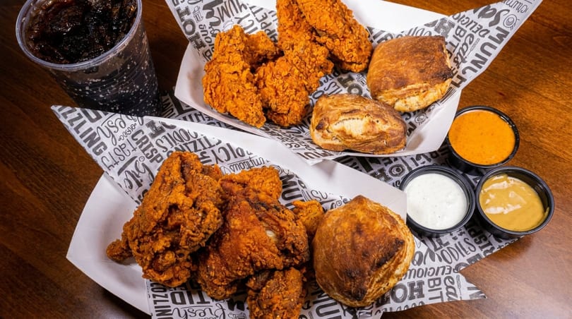 Two baskets of fried chicken and biscuits, with a soda and three dipping sauces on a wooden table.